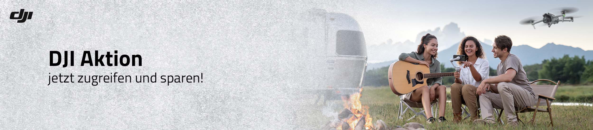 DJI Drohne im Flug über Campingplatz, drei Personen sitzen am Lagerfeuer mit Gitarre vor Wohnwagen