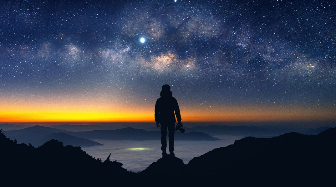 Milchstraße über Berglandschaft bei Nacht, Person als Silhouette auf Gipfel vor Sternenhimmel
