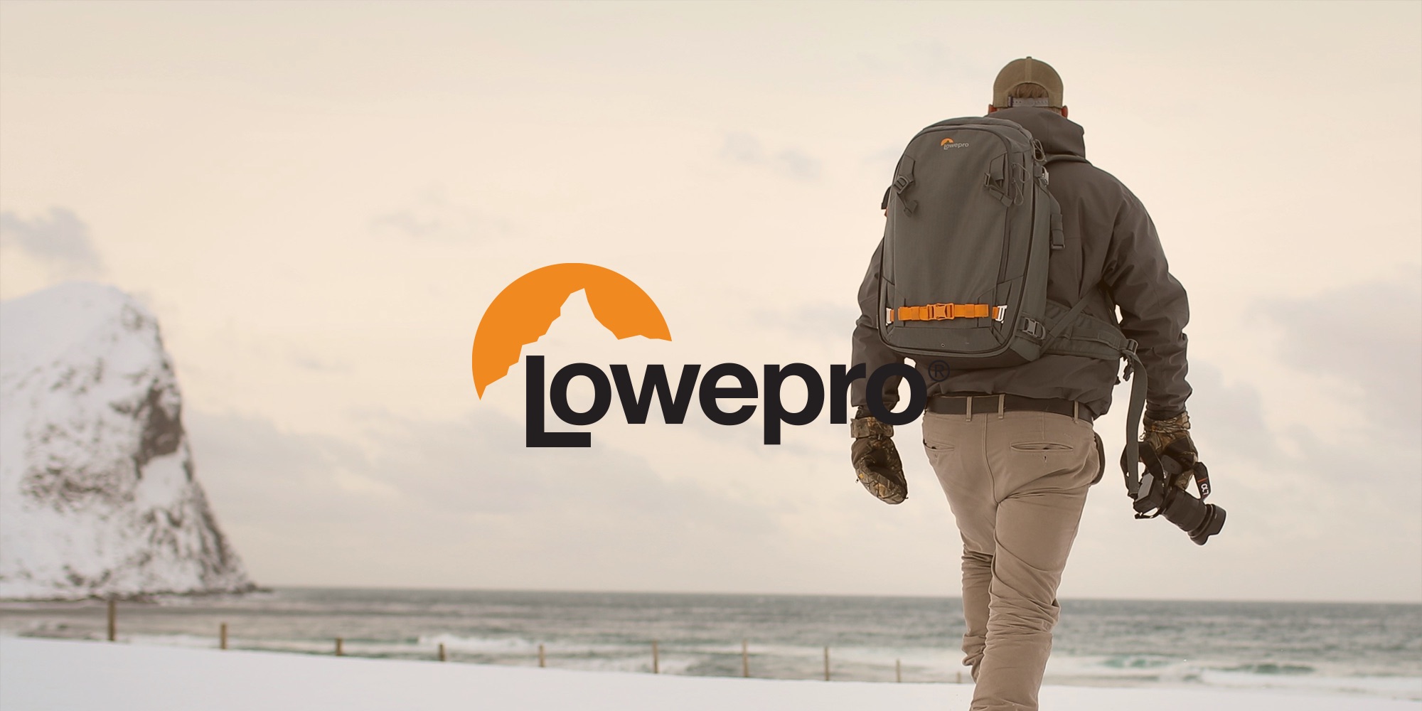 Fotograf mit Lowepro Fotorucksack auf verschneitem Strand, Kamera in der Hand, Blick Richtung Meer und Felsen
