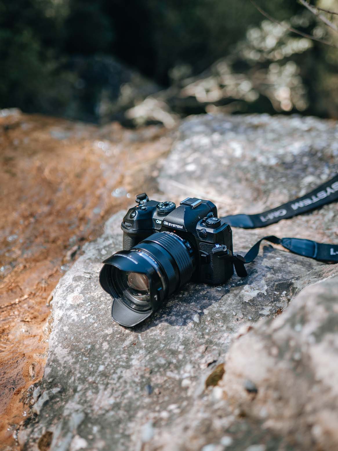 OM System Systemkamera mit Objektiv in schräger Ansicht auf einem Felsen neben fließendem Wasser platziert
