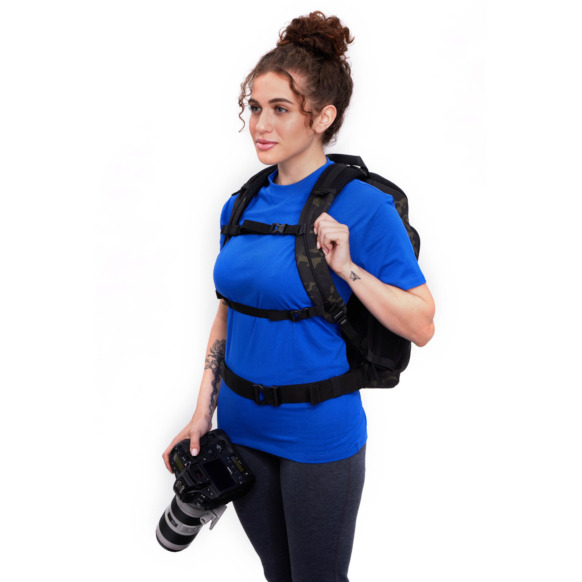 Axis V2 16L Rucksack (multicamo/schwarz) Tenba Axis V2 16L Rucksack in Multicamo Design, Lifestyle Foto mit einer Frau vor weissem Hintergrund und einer Kamera in der Hand