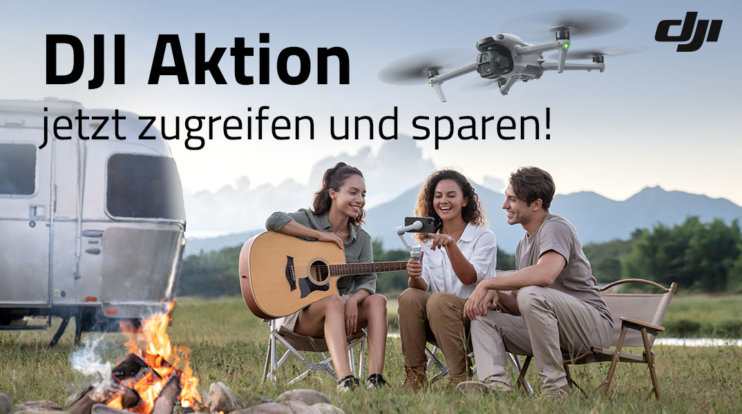 DJI Drohne in schräger Ansicht über Campingplatz, Gruppe sitzt vor Wohnwagen am Lagerfeuer mit Gitarre