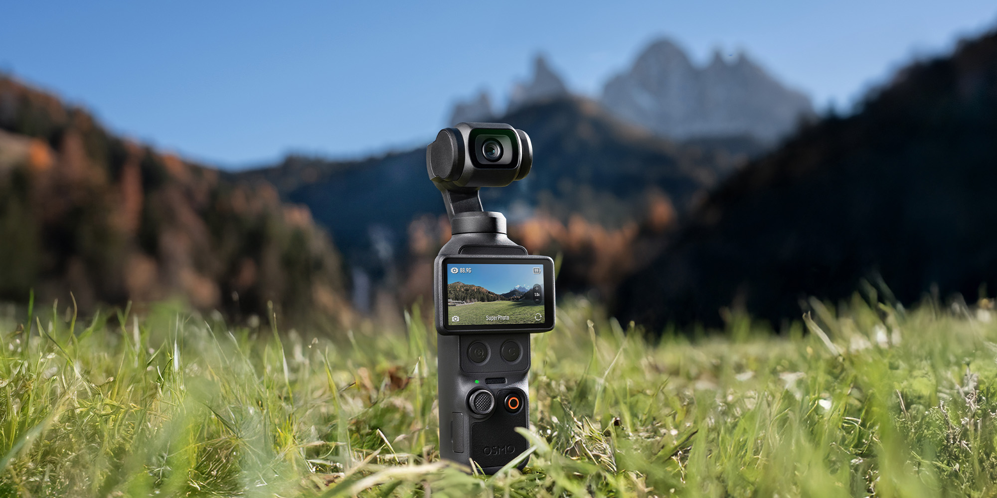 DJI Osmo Pocket 4 – Vorderansicht auf Wiese platziert, Display zeigt Landschaftsaufnahme vor Bergkulisse