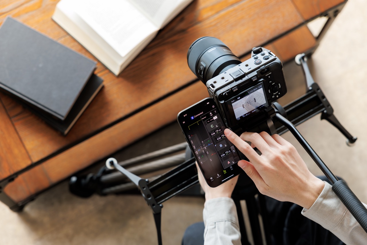 TAMRON Lens Utility auf Smartphone zur Fokussteuerung bei Kamera auf Schiene, Draufsicht auf Aufnahme-Setup mit Tischszene