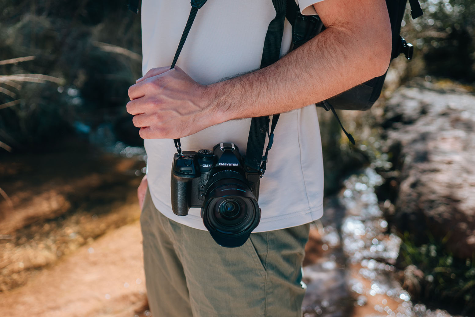 OM System OM-1 mit Objektiv frontal am Kameragurt getragen, aufgenommen bei einer Wanderung am Bach
