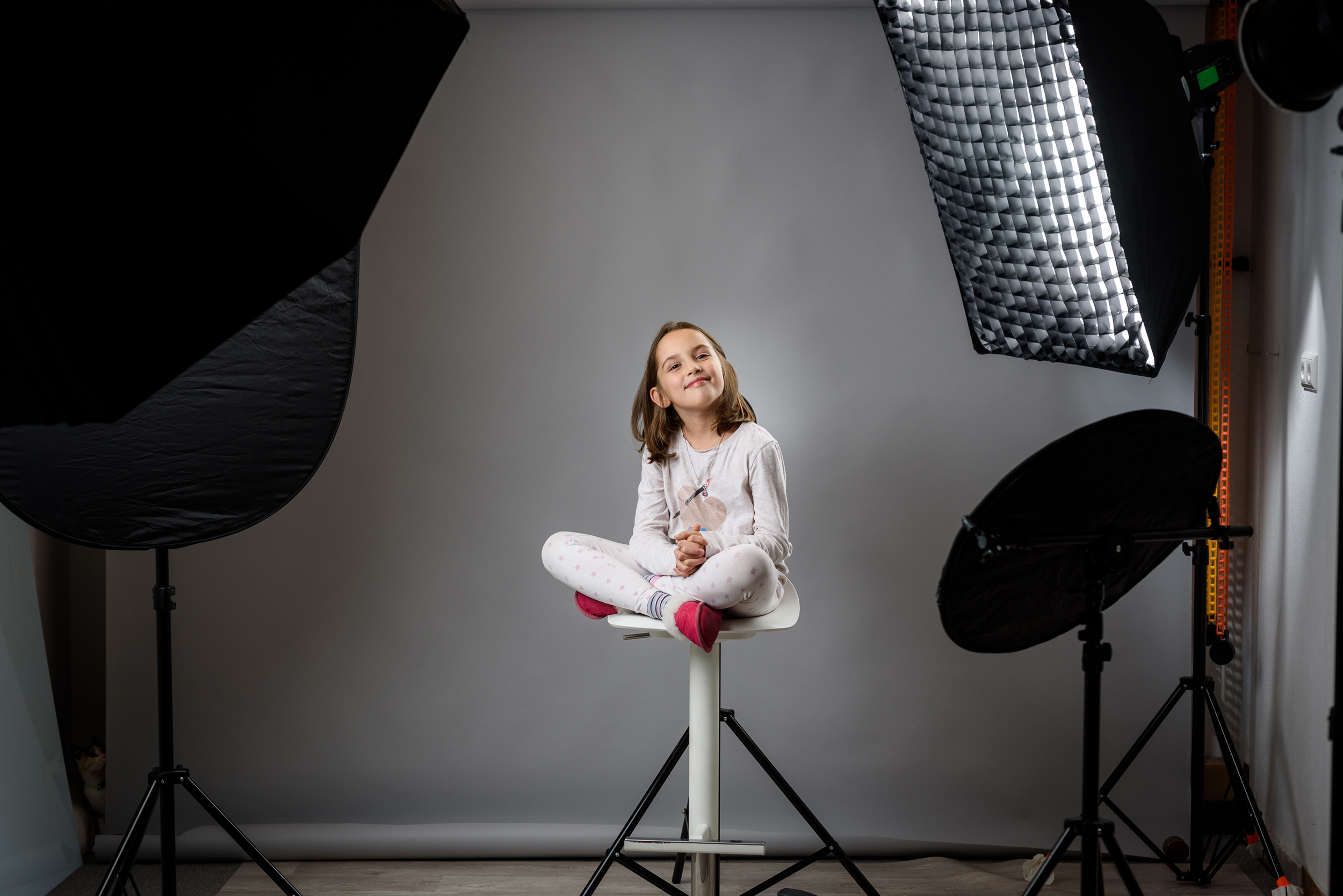 Kind sitzt vor grauem Hintergrund zwischen Softboxen und Reflektoren im Fotostudio