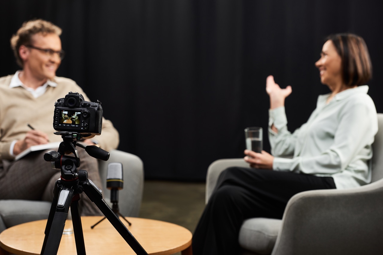 Interviewszene mit Kamera auf Stativ und zwei Personen im Gespräch vor schwarzem Hintergrund