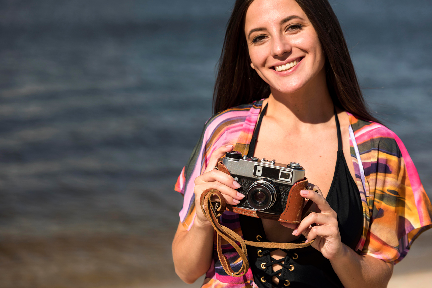 Reisefotografie – Frau am Strand hält eine analoge Kamera vor Meereskulisse