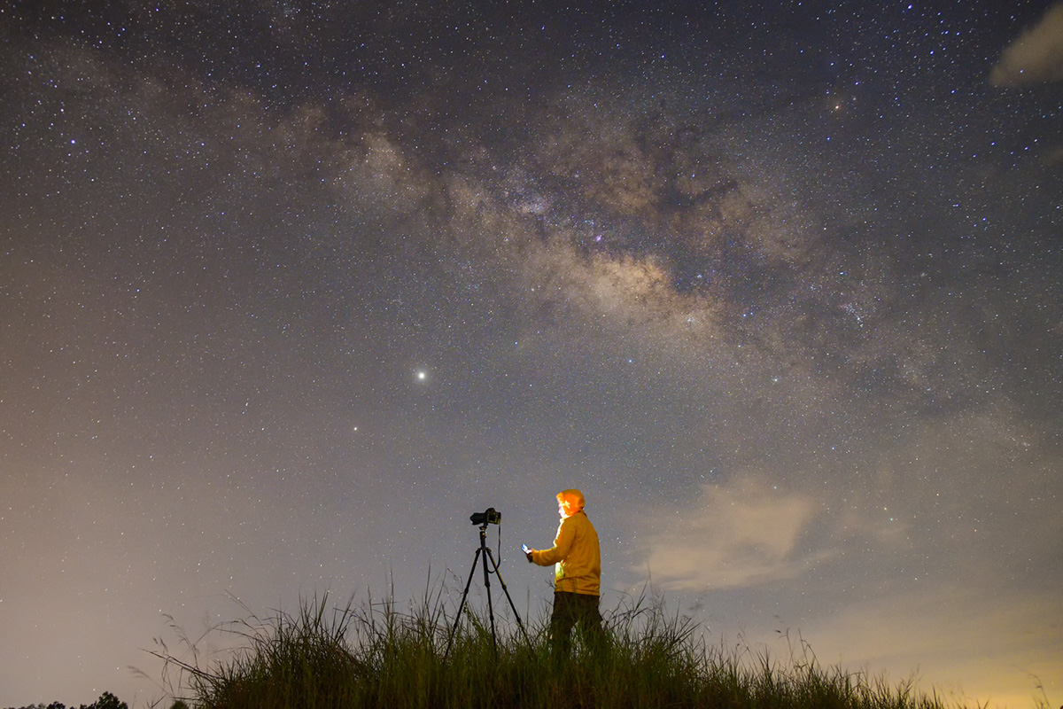 Fotograf mit Kamera auf Stativ unter Milchstraße, Nachtaufnahme mit sichtbaren Sternen und leichtem Wolkenzug