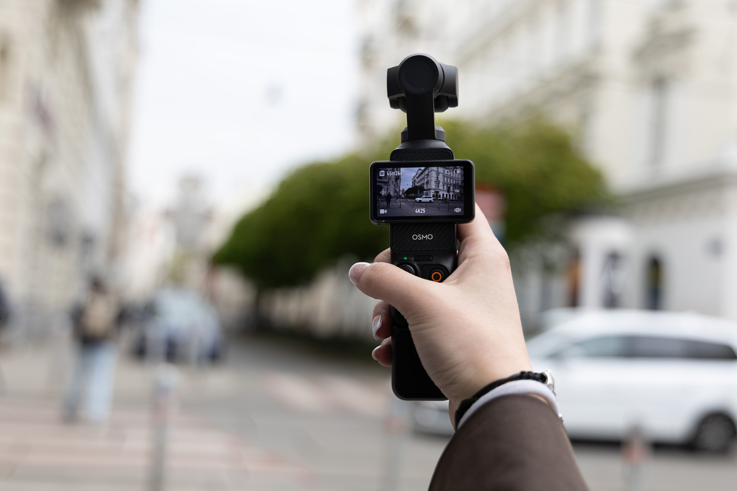 DJI Osmo Pocket 3 in Hand gehalten, Frontdisplay zeigt Aufnahme einer Straße mit Gebäuden im Hintergrund
